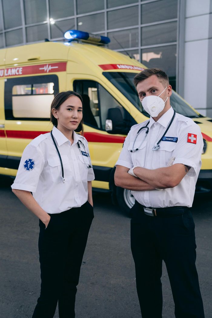 Two paramedics in uniform standing confidently beside an ambulance in an urban setting.